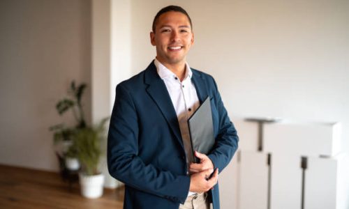 A mid-adult Hispanic male real estate agent smiles engagingly, clad in professional attire with a folder in hand, standing inside a modern office environment.